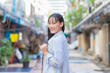 Confident young Asian female who wears a blue white shirt and bag smiles happily and looks at the camera as she commute to work through the old town.