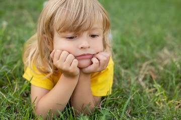 a blond boy with long hair and a mysterious thoughtful expression on his face lies on a green lawn