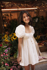 Naklejka premium Cute young caucasian woman smiles with her teeth at camera standing on backdrop of flower market. Brunette woman with wavy hair wears white sundress in summer. Lifestyle, different emotions, concept.