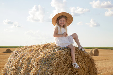 Naklejka na ściany i meble Portrait of cute little Caucasian girl in white dress sitting on hay stack on yellow wheat field