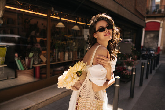 Pretty young caucasian girl posing looking at camera standing outdoors in sunny weather. Brunette woman with wavy hair wears casual summer clothes. Lifestyle concept