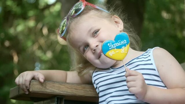 Little blonde girl holds gingerbread in Ukrainian national colors in her hand, it says "Ukraine is me" . Close-up portrait of girl sitting on park bench with cookies in ukrainian flag colors.  
