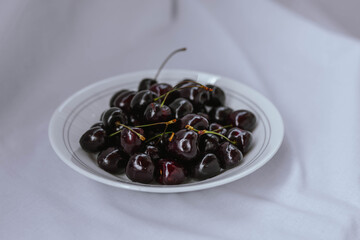Fresh ripe dark black huge sweet cherries and water drops on white plate, light cloth background. Rustic organic fruits to eat.  Bio and eco berries for jam, juice, smoothie, desserts. Selective focus