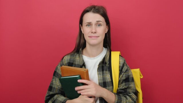 Confused young student woman 20s with yellow backpack holding notebooks making doubts gesture while lifting shoulders, posing isolated over red color background wall in studio. Education concept