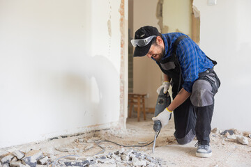 Removing a concrete floor with a hand hammer. Concrete rubble on the construction site of a house. Destruction of old tiles. Male construction worker fiercely working in overalls and protective mask.