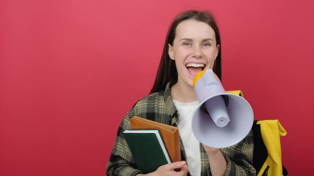 Portrait of promoter girl teen student 20s wears shirt and yellow backpack holding books scream in megaphone announces discounts sale Hurry up, posing isolated over red color background in studio