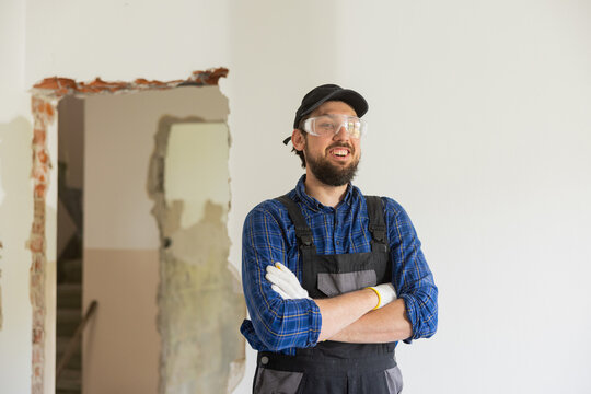 Smiling man in baseball cap wearing protective suit and plaid shirt has goggles on to protect eyes hands crossed over chest stands in middle of renovated house.