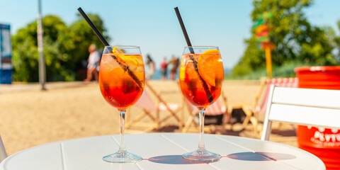 Banner,advertisement.Two glasses of orange spritz aperol drink cocktail on table outdoors with sea and trees view blurred background.
