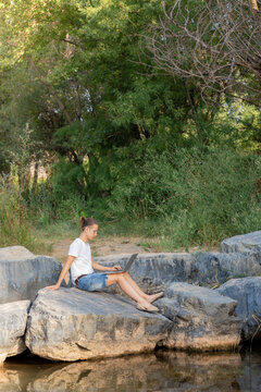 Vertical Vision Of A Remote Worker Sitting On The Bank Of A River And Working With His Laptop In Nature