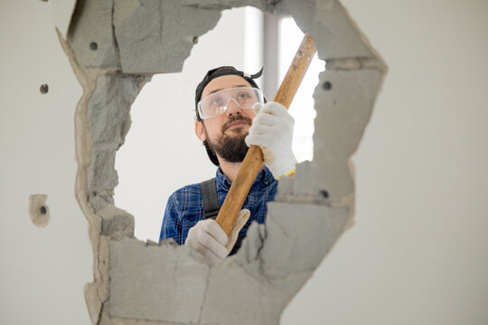 A smiling guy demolishes a wall in a house using a construction hammer on a long stick. Fun demolition of an apartment during renovation, getting rid of an unnecessary wall gaining space.
