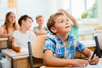 Cute happy blond school boy sitting and listens carefully to the teacher in primary class. Smiling n child pupil studying and writing at school.