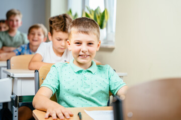 Smmilling little boy sitting at desk in class room and looking at camera. Portrait of young caucasian schoolboy studying with classmates in background.