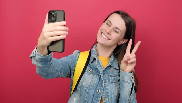 Portrait of young woman student in denim jacket and yellow backpack do selfie shot mobile cell phone, posing isolated on plain red background wall. Education in high school university college concept
