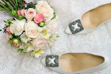 Wedding day composition with silk white shoes, pastel bridal bouquet on white lace blurry background. Top view