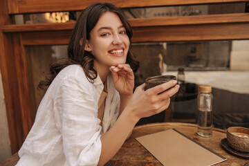 Cheerful young caucasian lady wearing shirt spends leisure time drinking cup of coffee in street cafe during day. Brunette with wavy hair is looking at camera. Concept of enjoying weekend, vacation