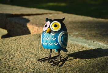 A close-up toy of a surprised blue owl with multi-colored eyes stands on a granite stone. The toy is illuminated by evening sunlight and casts a shadow