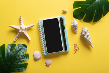 A smartphone, glasses, a blank notebook and Monstera Deliciosa leaves and shells on a yellow background. Blogging in the summer at the workplace is a business