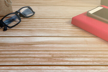 Cellphone, red note book, potted plant and reading glasses on wooden desk.