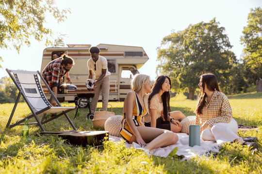 A group of friends spend time together in nature. The girls are sitting on a blanket and talking. The boys are preparing a barbecue