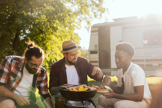 A group of friends spend time together in nature. The middle-aged men prepare a barbecue near the RV. A bearded boy carries snacks for the barbecue.
