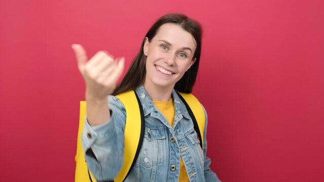 Happy young student woman wearing yellow backpack and denim jacket beckoning come here gesture with hand inviting welcoming happy and smiling, posing isolated over red color background wall in studio