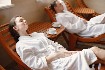 Cute couple in white bathrobes laying on a sunbeds indoors and drink a tea