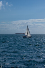Maritime landscape with a sailboat and an island. A sailboat with a white sail sails in a small sea, behind we can see an island and a signaling buoy. It is the open sea.