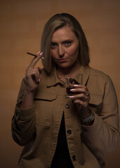 Obraz premium Portrait of a young, beautiful girl with blue eyes and blonde hair in a beige jacket. She looks sternly, smokes a cigarette and holds a glass of whiskey. Expensive watches, gold chain and rings.