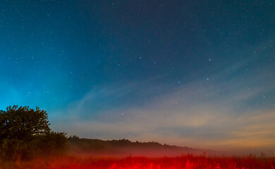 Fototapeta premium The night sky with many bright stars over the forest and the field with a red glow. A nighttime abstract landscape. 