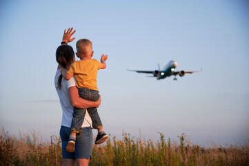 Back view of woman with child waving hands to flying commercial airplane in the evening. Lifestyle...
