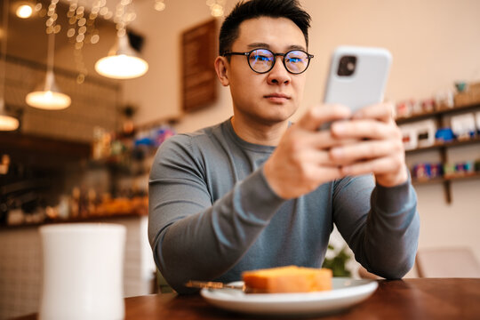 Adult asian man using cellphone while having breakfast in cafe indoors