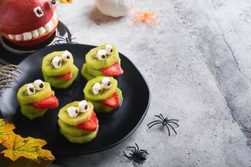 Spooky green kiwi monsters for Halloween. Healthy Fruit Halloween Treats. Halloween party kiwi, strawberry, apple and marshmallow monster on grey stone or concrete table background. Selective focus.