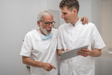 Mature smiling doctor and young intern doctor discussing patient diagnosis, holding digital tablet. Father and son dentist doctors