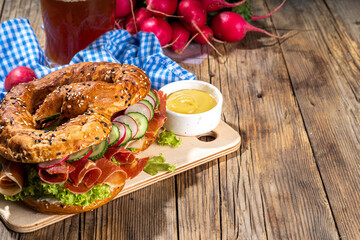 Oktoberfest pretzel bagel sandwich. Oktoberfest day breakfast, burger recipe for holiday beer party. Big pretzel with ham, cheese, fresh vegetables, with radish, beer, checkered tablecloth copy space