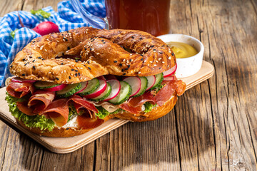 Oktoberfest pretzel bagel sandwich. Oktoberfest day breakfast, burger recipe for holiday beer party. Big pretzel with ham, cheese, fresh vegetables, with radish, beer, checkered tablecloth copy space