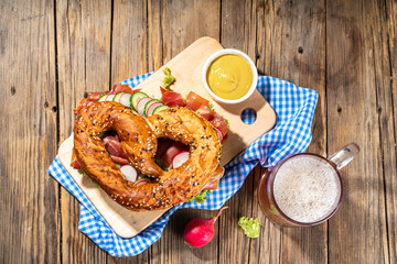 Oktoberfest pretzel bagel sandwich. Oktoberfest day breakfast, burger recipe for holiday beer party. Big pretzel with ham, cheese, fresh vegetables, with radish, beer, checkered tablecloth copy space