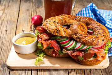 Oktoberfest pretzel bagel sandwich. Oktoberfest day breakfast, burger recipe for holiday beer party. Big pretzel with ham, cheese, fresh vegetables, with radish, beer, checkered tablecloth copy space