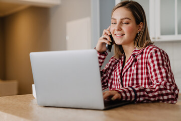 Young happy woman talking on cellphone while using laptop at home