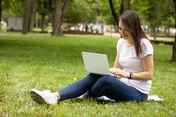 Portrait of pretty young woman sitting on green grass in park with laptop. Happy woman, student, freelancer with wireless headphones calling on laptop, talk by webcam, video conference.