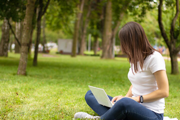 Young female side view. Woman working on laptop computer with blank black blank screen to copy space in city park on green grass on sunny lawn outdoors. Mobile office. Freelance business concept