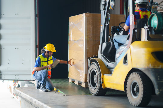 Asian forklift driver loading a shipping cargo container with a full pallet with boxes in logistics port terminal. Asian warehouse worker and safety inspector with digital tablet manage the process.