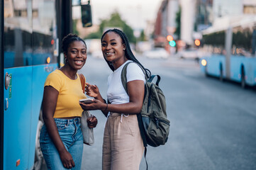 Two african american woman waiting for a bus and using a smartphone