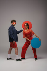 full length of stylish couple in blue and red outfits holding round shape carton on grey.