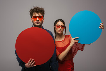 young couple in blue and red outfits and sunglasses holding round shape carton isolated on grey.