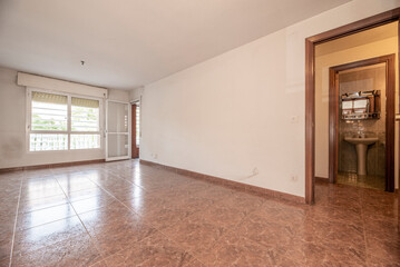 Empty living room with reddish stoneware floor, white painted walls, windows and exit door to a...