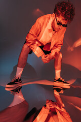 full length of curly man in sunglasses looking at reflection on grey with colorful light.