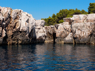 Beautiful view of the rocky coast of the island with a forest and blue sky in the background, a view from the sea - obrazy, fototapety, plakaty