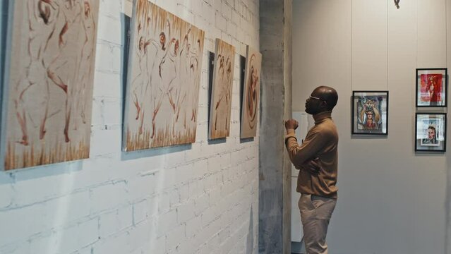 Medium long of young intelligent Black man observing collection of paintings on wall of modern art gallery