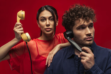 young woman and curly man holding retro handsets while posing on red.