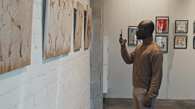 Medium long of young intelligent Black man taking pictures with smartphone of series of paintings on wall of modern art gallery